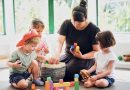 A mother playing calmly with her toddlers using colorful wooden blocks on the floor.