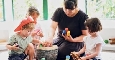 A mother playing calmly with her toddlers using colorful wooden blocks on the floor.