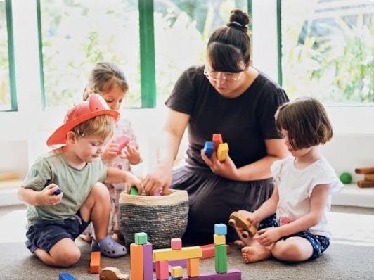 A mother playing calmly with her toddlers using colorful wooden blocks on the floor.